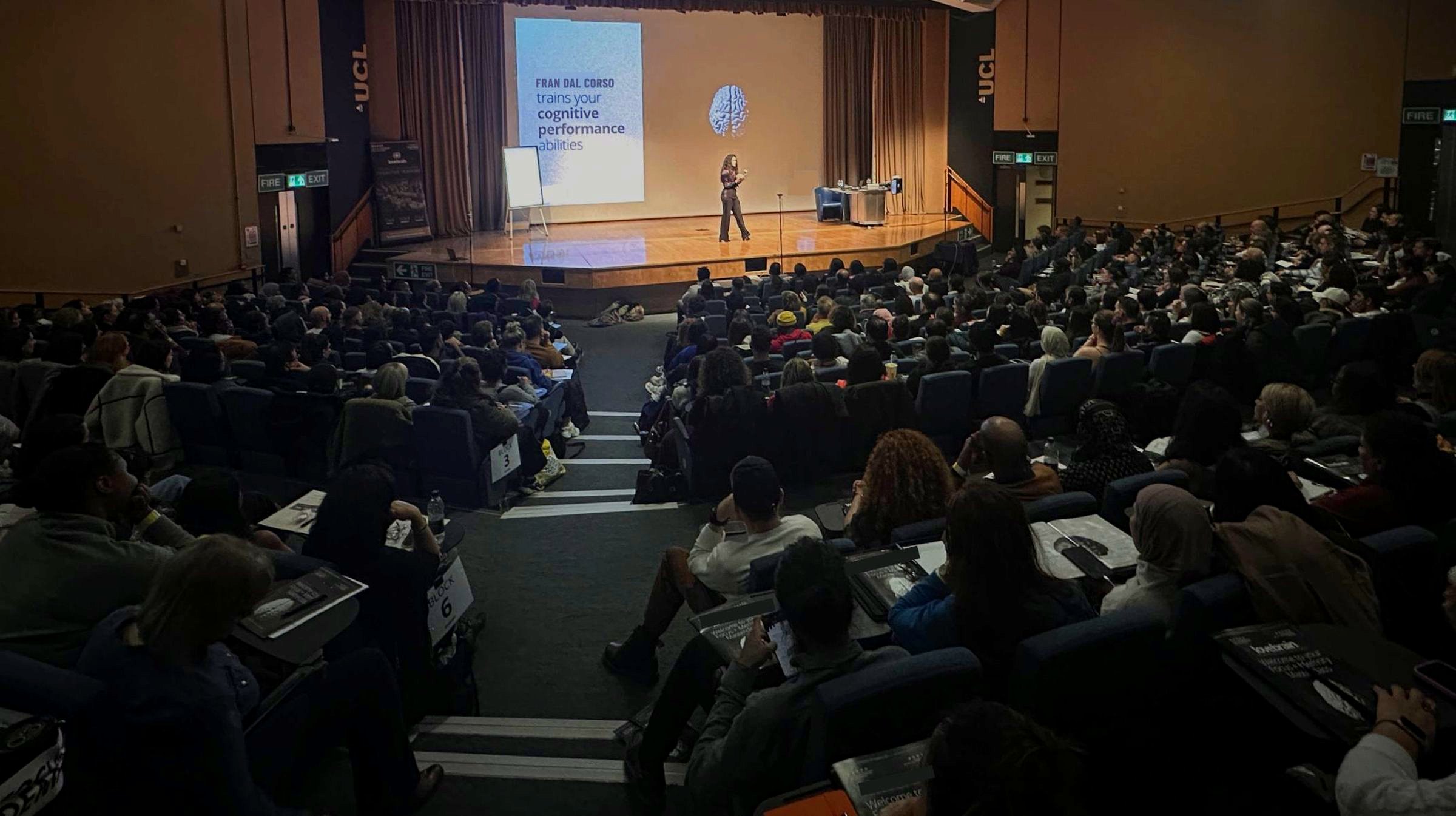 Fran Dal Corso on stage in front of a packed UCL lecture hall, audience taking notes
